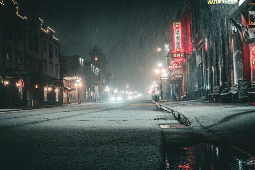 A snow-covered street at night with illuminated buildings and a river in the foreground. 5
