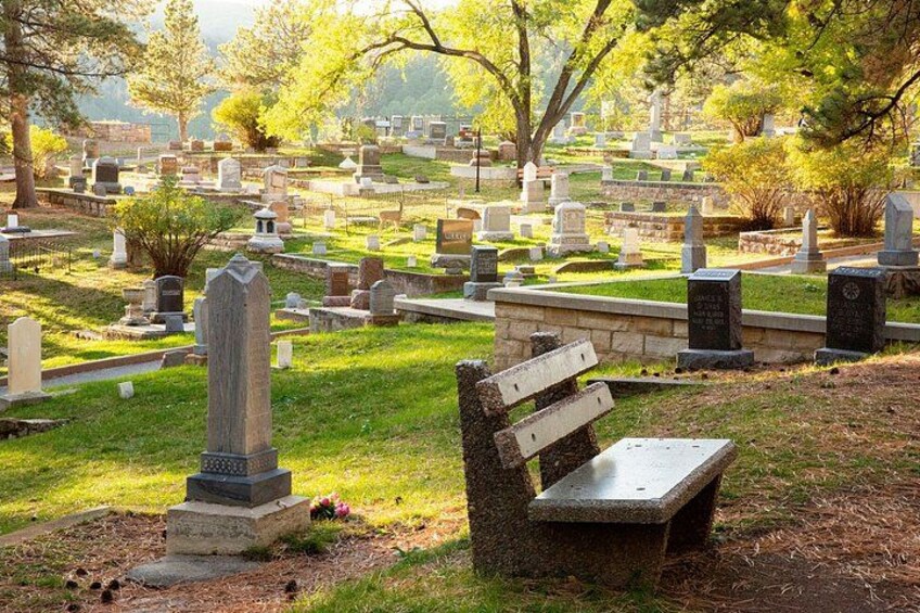 A cemetery with numerous headstones, a wooden bench, and trees providing shade. 5