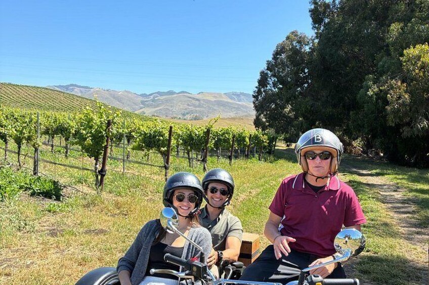 Three people on a motorcycle in a vineyard with rows of grapevines and hills in the background. 3