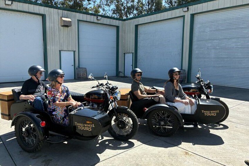Four people on motorcycles with sidecars parked in front of industrial buildings. 2