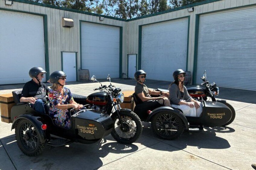 Four people on motorcycles with sidecars parked in front of industrial buildings. 2
