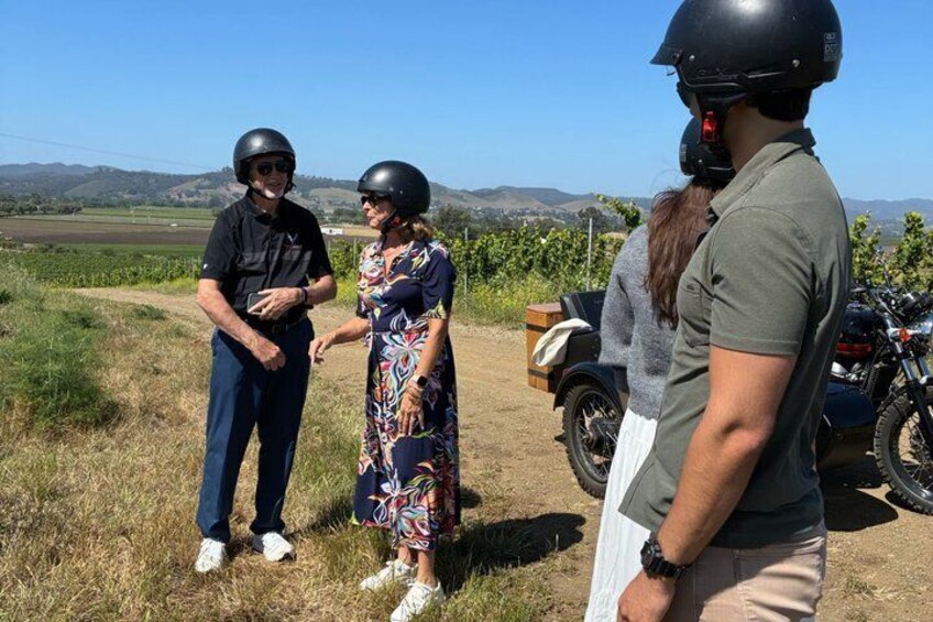 Three people standing on a dirt path with a motorcycle and a box nearby, surrounded by a rural landscape with fields and hills. 5