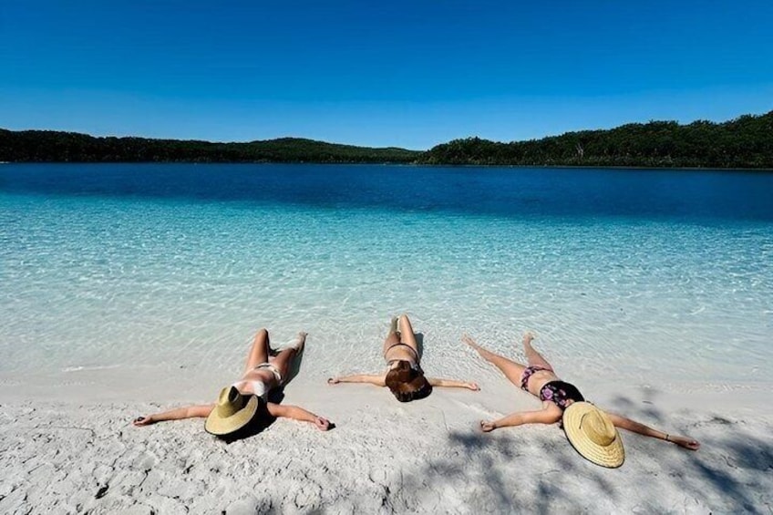 Three people lying on a beach with clear turquoise water and a forested island in the background. 3