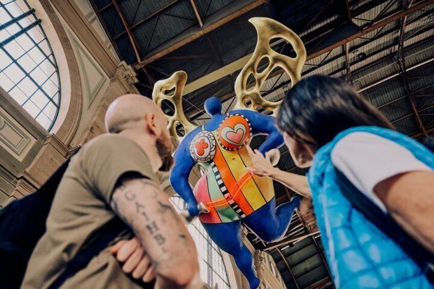 A person in a blue costume with a heart-shaped prop in a large indoor space with high ceilings and metal beams. 3