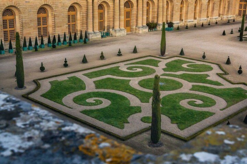 A courtyard with a geometric garden design, a building with arched windows, and a row of conical trees. 4