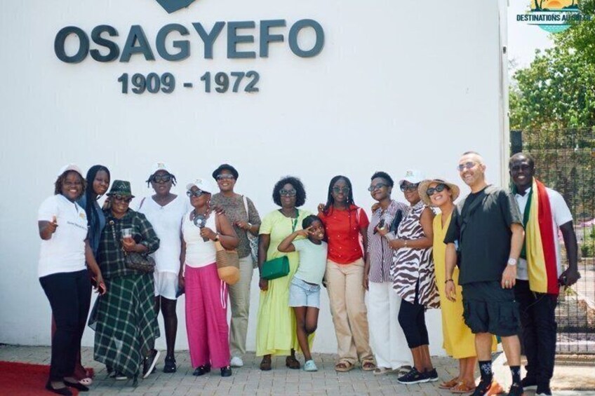 A group of people posing in front of a building with a sign reading 'OSAGYEFO 1909-1972.' 1