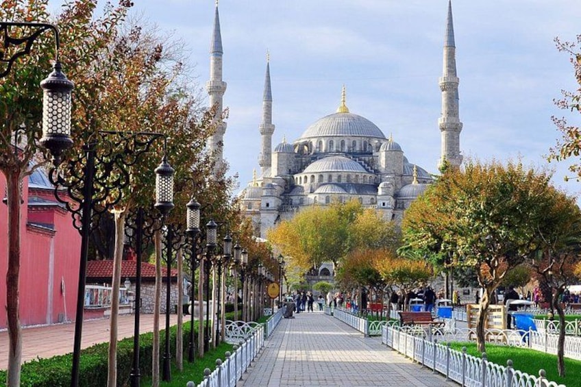 A mosque with multiple domes and minarets, a paved walkway lined with trees and street lamps, and a red building on the left. 2