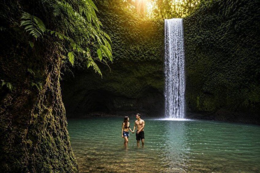 A waterfall in a forested area with clear water and two people standing in the foreground. 4