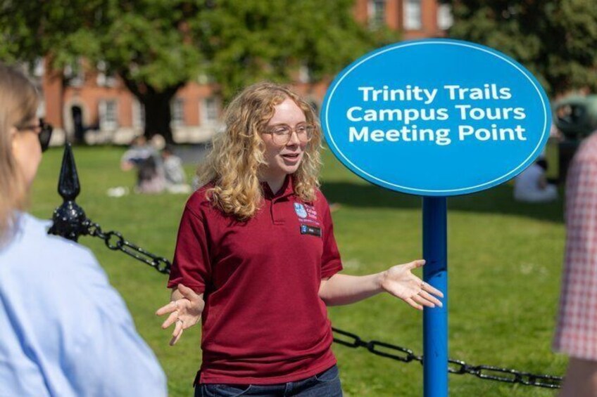 A person in a maroon shirt standing next to a sign that reads 'Trinity Trails Campus Tours Meeting Point.' 1