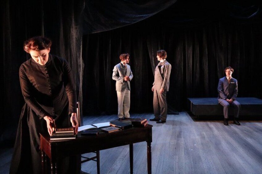 A stage with a person reading from a book, three individuals standing, and one seated on a bench. 2