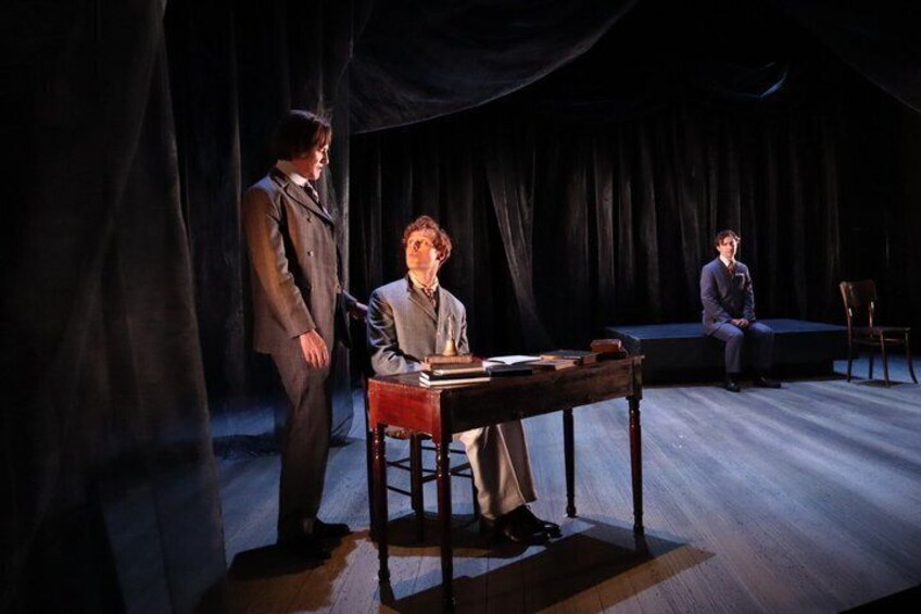 A stage with three people, one standing and two seated, with a table and chairs in the foreground. 5
