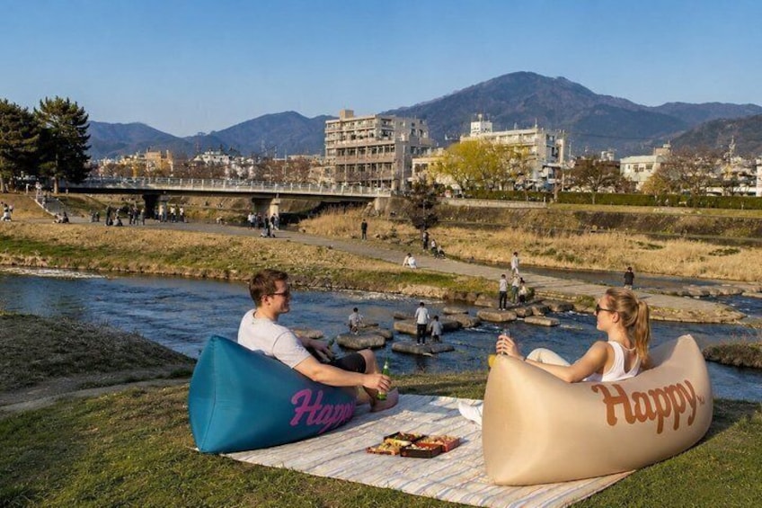 Two people sitting on bean bags by a river with a bridge and buildings in the background. 2