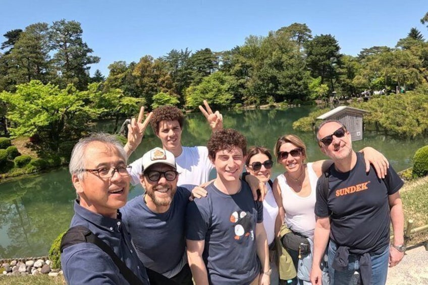 A group of people posing by a lake with trees and a small building in the background. 1