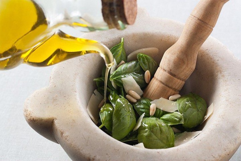 A mortar with spinach and a wooden pestle, olive oil being poured into it. 1