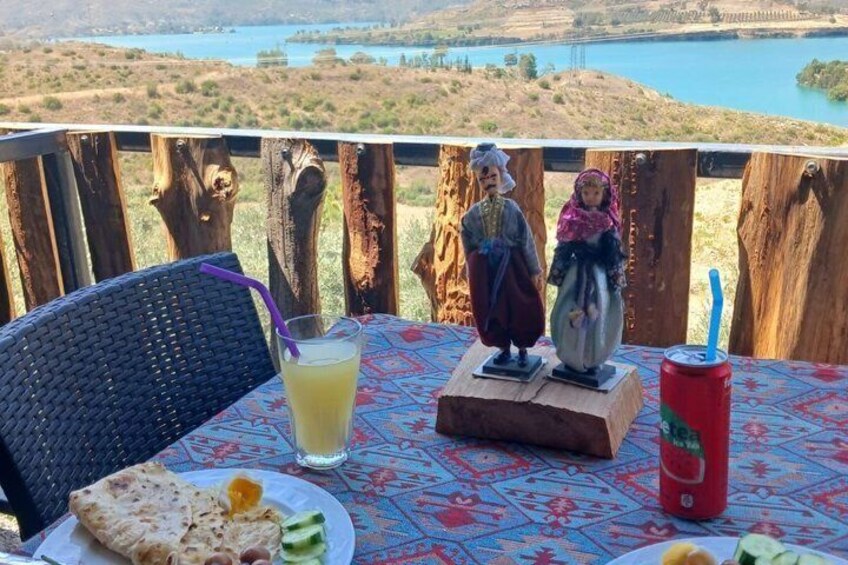 A table with a drink, a plate with food, and a figurine, overlooking a lake and trees. 3