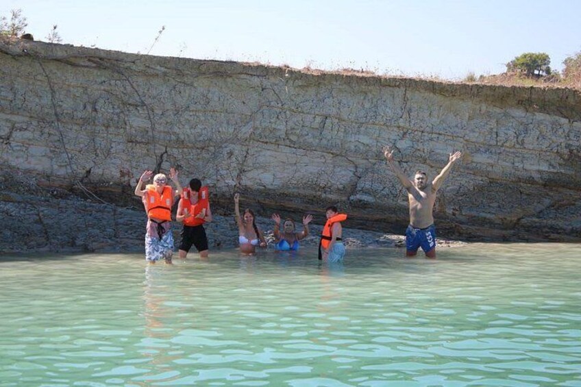 People in a shallow water body, some wearing life jackets, with a rocky cliff in the background. 2