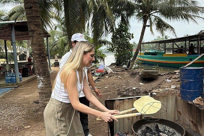A person cooking outdoors with a large pot over an open fire, palm trees, and a boat in the background. 3