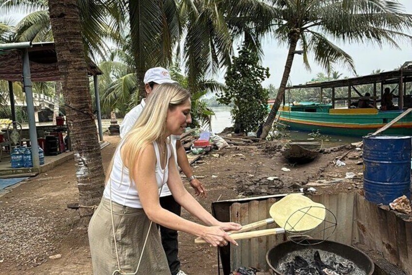 A person cooking outdoors with a large pan on a stove, palm trees, and a boat in the background. 3