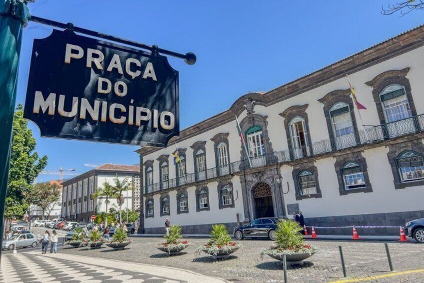 A historic building with a sign reading 'Praça do Município' and a paved plaza with planters. 5