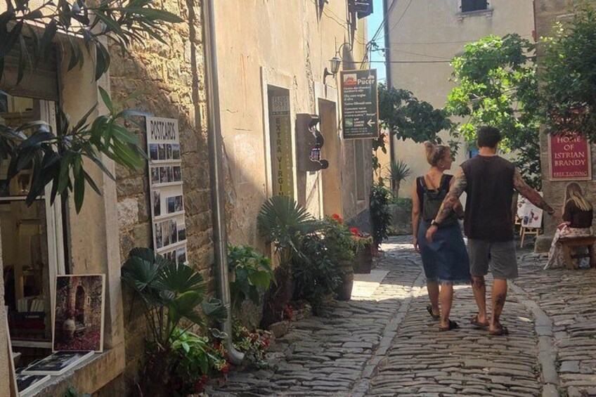 A cobblestone street with a postcard display, a sign for 'House of Istrian Frandly,' and a café with outdoor seating. 2