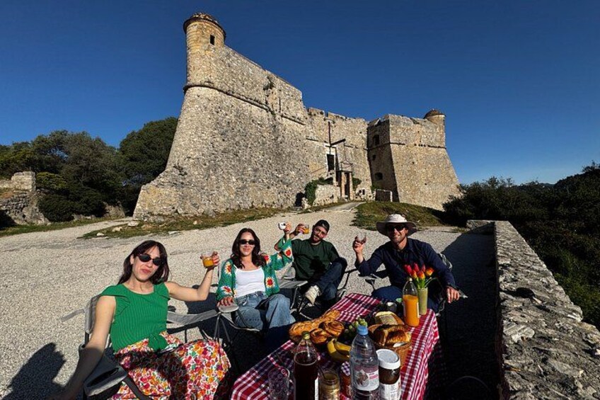 A group of people sitting at a table with food and drinks, with a historic stone building in the background. 3