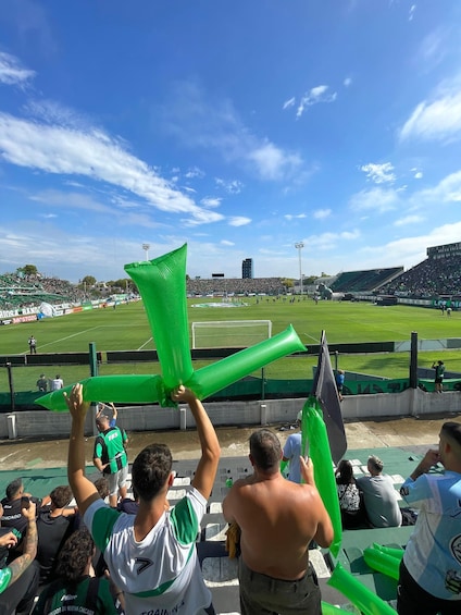 A stadium with spectators holding green inflatable tubes, a soccer field, and a clear blue sky. 4