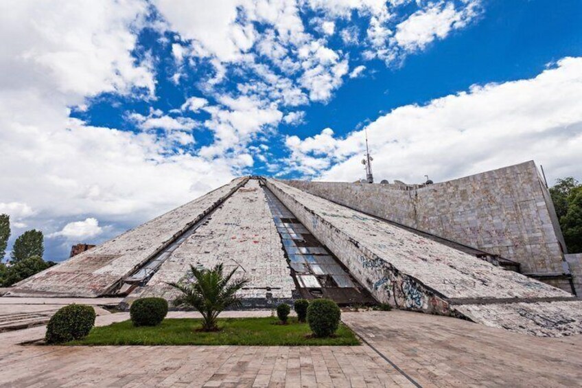 A pyramid-shaped monument with graffiti, surrounded by a paved area and small trees. 2