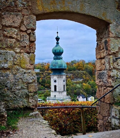 A stone archway framing a tower with a green dome and clock, set against a backdrop of autumn foliage and houses. 5