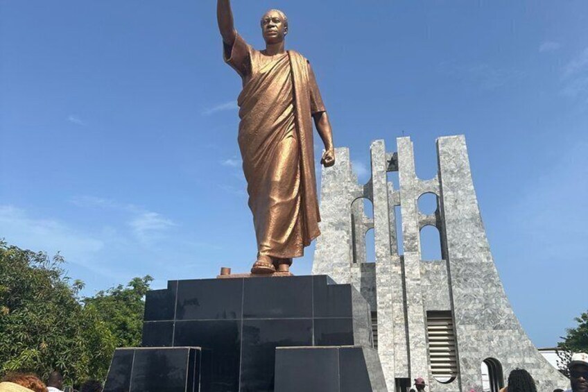 A statue of a person in a robe standing on a pedestal with a large, modern structure behind it. 1