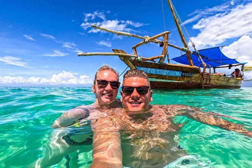 Two people swimming in clear turquoise water with a traditional boat in the background. 1