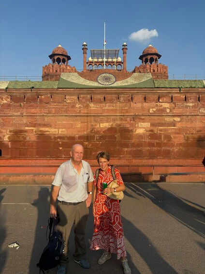 A man and woman standing in front of a large red brick wall with a green roof and a flagpole. 5