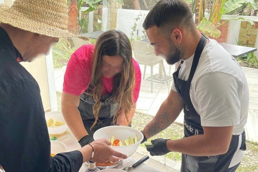 Three people preparing food outdoors, with a table and a bowl of ingredients. 2