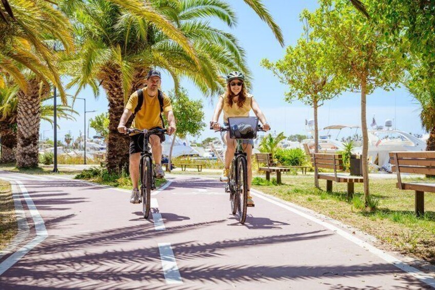 Two cyclists on a paved path with palm trees and benches in a sunny outdoor setting. 1