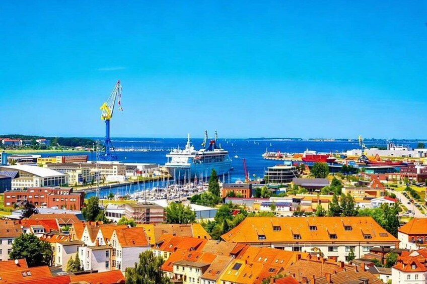 A coastal cityscape with a prominent shipyard, a marina with boats, and historic buildings with red-tiled roofs. 1