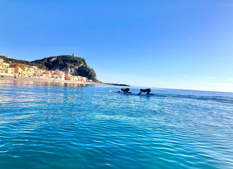 A coastal town with buildings along the shore and two people in the water. 1
