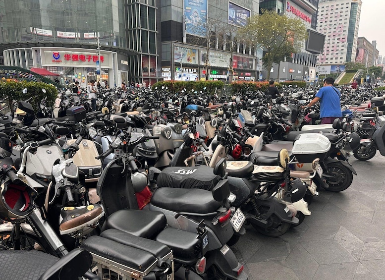 A large number of parked scooters and motorcycles in an urban setting with commercial buildings in the background. 5