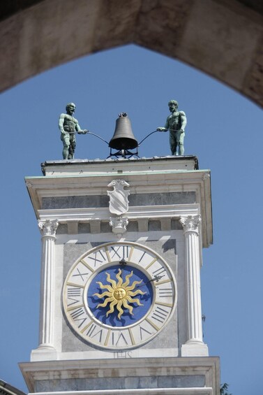 A clock tower with a sun motif, two bronze statues, and a bell. 1