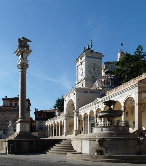 A historic square with a clock tower, a fountain, and classical architecture. 4