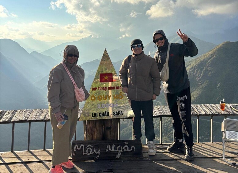 Three people standing near a wooden sign with a mountain view in the background. 1
