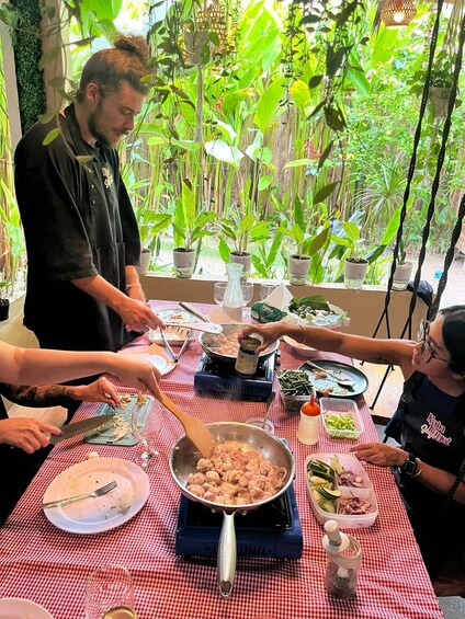 A group of people cooking outdoors with a checkered tablecloth, surrounded by lush greenery. 3