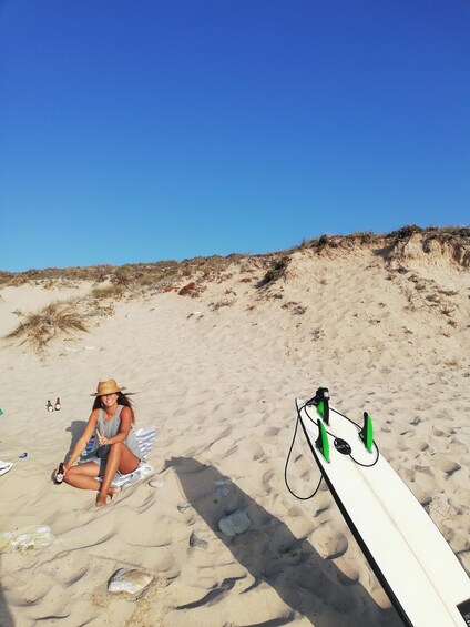 A person sitting on a beach with a surfboard leaning against the sand. 2