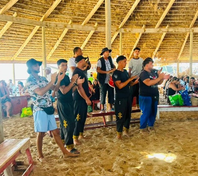 A group of people standing on a sandy beach under a thatched roof structure. 2