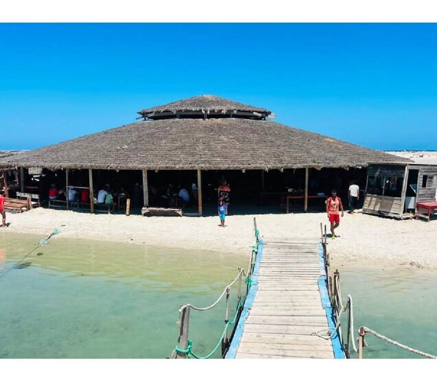 A beachside structure with a thatched roof, a wooden dock leading to it, and people walking on the sand. 3