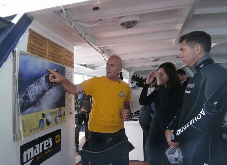 A man pointing at a poster with a shipwreck illustration, a woman looking at a poster, and a man in a black jacket with 'Mare's' on it. 1
