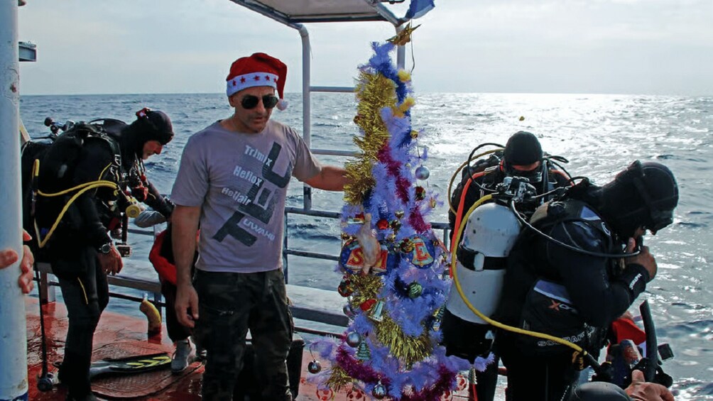 A person wearing a Santa hat standing on a boat with scuba divers and a decorated Christmas tree. 5