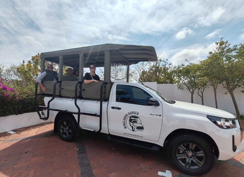 A white pickup truck with a roof rack and passengers, parked on a brick pavement with trees and a white wall in the background. 5