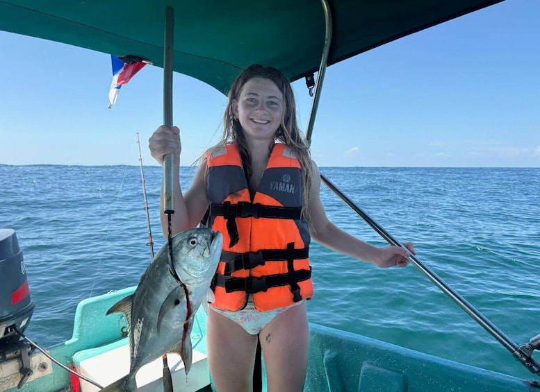 A person on a boat holding a fish, wearing a life jacket, with a flag and calm sea in the background. 5