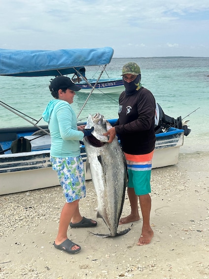 Two people holding a large fish on a beach with a boat in the background. 1