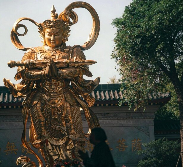A golden statue of a deity with elaborate armor and a curved headdress, set against a traditional building with a tiled roof. 3
