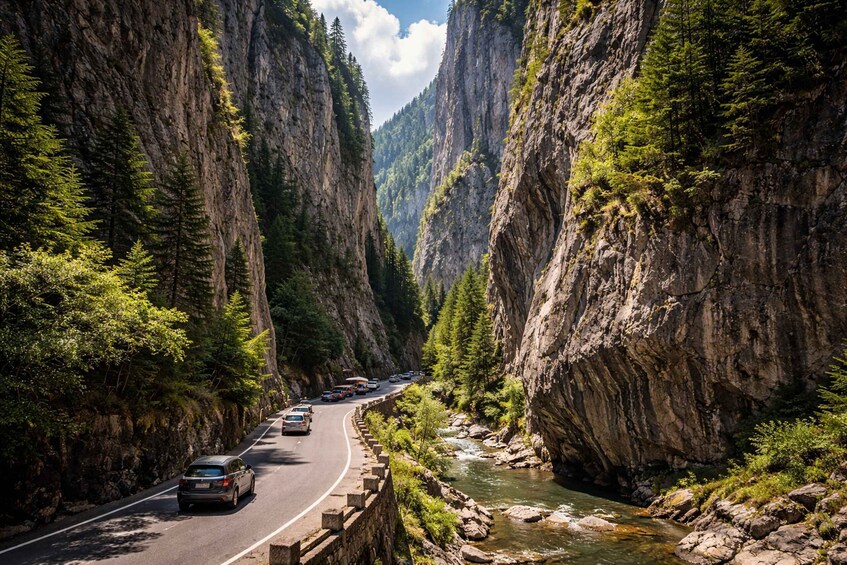 A road flanked by steep cliffs and a river, with cars driving along the road. 1